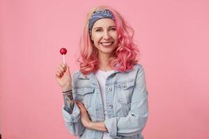 Young positive beautiful pink haired lady in denim shir, holding a lollipop, looks at the camera and broadly smiling, stands over pink background. photo