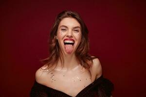 Joyful pretty young brown haired woman with festive makeup wearing elegant black top with red dots while posing over claret background, having fun and showing happily tongue photo