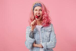 Young agressive beautiful pink haired woman with one eye closed in denim shir, holding and trying to bite off lollipop, stands over pink background. photo