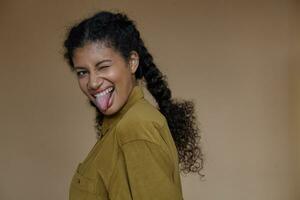 Happy lovely young curly brunette dark skinned woman with braided hairstyle having fun while posing over beige background, winking cheerfully to camera and showing out tongue photo