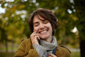 Pleasant looking young cheerful lovely short haired female keeping her eyes closed while smiling happily during nice phone talk, standing over park background in warm cosy wear photo