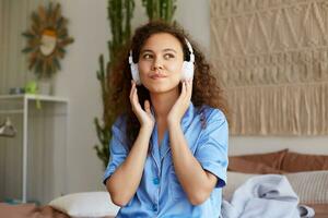 foto de soñando Rizado joven bonito africano americano dama, escuchando favorito canción en auriculares, mira lejos ingenio contento expresión.