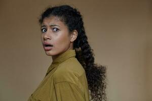 Portrait of frightened young dark skinned woman wearing her curly brown hair braided while standing against beige background, looking scaredly to camera and frowning her eyebrows photo