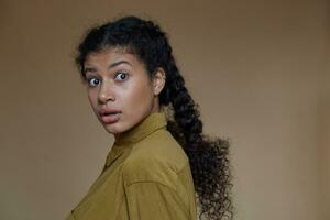 Amazed pretty young curly brunette female with dark skin looking at camera with wide eyes opened, keeping her long hair in braids while posing over beige background in mustard shirt photo