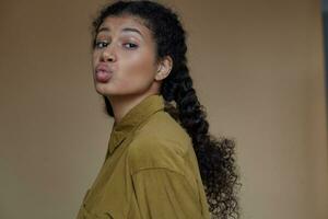 Close-up of positive lovely young dark skinned woman wearing her curly brown hair braided, folding lips in air kiss while looking at camera, dressed in casual clothes over beige background photo