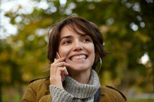 Portraitf of beautiful glad young brunette woman with short haircut keeping mobile phone in raised hand while having pleasant talk, walking through city garden and smiling broadly photo