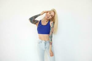 Pleasant looking young tattooed blonde woman with casual hairstyle smiling nicely with closed eyes while enjoying music track, standing over white background photo