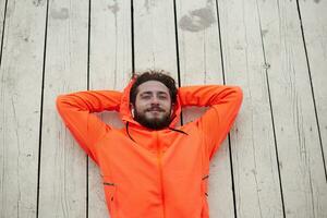 Pleased young brown haired bearded man folding raised hands under his head while lying on wooden floor, listening to favourite song in his earphones and smiling positively photo