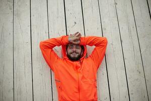 Photo of charming young sporty brunette man with beard smiling cheerfully and keeping hands on his forehead, lying on outdoor wooden floor after morning running session