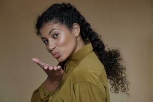 Attractive young curly brunette lady with dark skin wearing braided hairstyle while standing over beige background, raising palm and blowing air kiss to camera photo