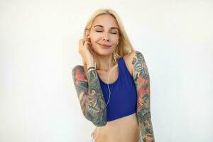 estudio Disparo de joven positivo tatuado mujer con casual peinado sonriente agradablemente con cerrado ojos mientras escuchando a música en su auriculares, posando terminado blanco antecedentes foto