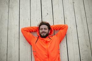 Cheerful young dark haired bearded man with earphones keeping his head on raised hands while lying over outdoor terrace and looking happily aside with charming smile, dressed in sporty wear photo