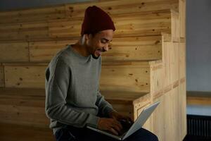 Indoor shot of cheerful pretty dark skinned male with beard sitting over stairs and keeping laptop, looking at screen joyfully and holding hands on keyboard photo