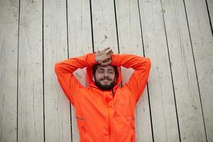 Portrait of positive pretty young bearded sportsman wearing piercing in his eyebrow, relaxing on wooden floor after hard workout outdoor, dressed in warm sporty orange hoodie photo