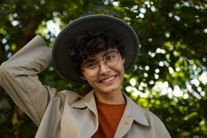 Shot of cheerful brunette pretty woman with curly short hair wearing trendy outfit and eyewear while posing over green city palk on sunny day, smiling to camera positively and keeping hand on her hat photo