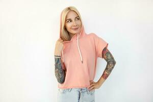 Positive young beautiful tattooed blonde woman dressed in sporty clothes keeping her hand on waist while looking gladly with pleasant smile, isolated over white background photo