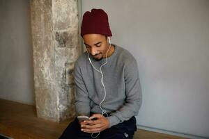 Good looking young bearded dark skinned guy sitting on long wooden bench with headphones and mobile phone, chatting with friends, dressed in grey sweater and burgundy headdress photo