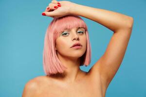 Studio photo of charming young green-eyed female with short pink haircut keeping raised hand under her head and looking calmly at camera, isolated over blue background