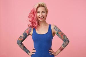 Portrait of happy cute lady with pink hair and tattooed hands, standing over pink background and broadly smiling, wearing a blue shirt. People and emoyion concept. photo