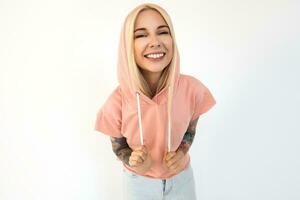 Portrait of pleased young tattooed blonde woman with nose piercing looking happily at camera with wide smile while standing over white background in sporty wear photo