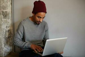 Good looking young bearded freelancer with dark skin preparing materials for presentation on his laptop, keeping hands on keyboard and looking on screen attentively photo