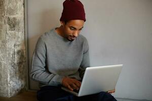 Portrait of handsome bearded dark skinned man dressed in grey sweater and burgundy cap working out of office with his modern laptop and typing letter with keyboard photo