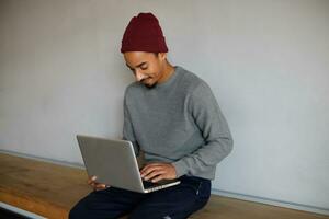 Pleasant looking young bearded dark skinned man in grey sweater, blue trousers and burgundy cap sitting over white wall and chatting with his friends on his laptop photo