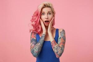 Portrait of young wondered nice pink haired lady, with palms on cheeks, hears unbelievable news, looks at the camera with wide open mouth in surprise, stands over pink background. photo