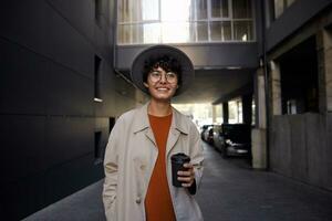 Lifestyle fashion photo of young pretty female with short curly hair walking over city parking and keeping takeaway coffee in her hand, wearing trendy outfit and smiling sincerely