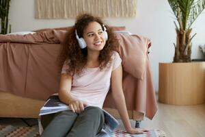 retrato de alegre joven Rizado mulato dama sentado en habitación, disfrutando su favorito canción y leyendo nuevo revista acerca de arte, pensativamente mira lejos y gasto bueno hora a hogar. foto