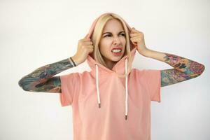 Displeased young attractive blonde lady with tattoos holding her hood with raised hands and grimacing her face while looking aside, isolated over white background photo