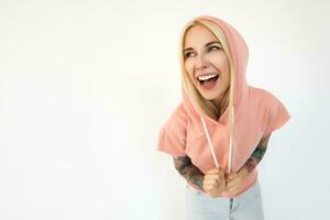 Indoor photo of young cheerful tattooed blonde female with natural makeup laughing happily while wearing hood of her t-shirt, isolated over white background