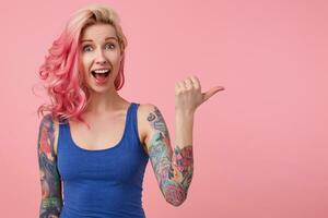 amazed nice pink haired lady in blue t-shirt, hears unbelievable news, with wide open mouth in surprise, broadly smiling, stands over pink background and pointing to the right at copy space. photo