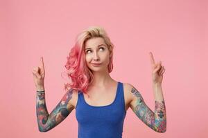 Young beautiful pink haired lady in blue t-shirt with arms raised, doubts something, looks up and wants to draw your attention pointing with fingers on the copy space, standing over pink background. photo