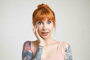 Portrait of young pretty surprised redhead lady with nose piercing keeping palm on her cheek while looking amazedly at camera, isolated over white background photo