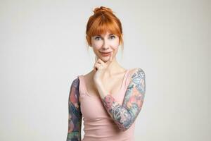 Cheerful young attractive redhead woman with bun hairstyle keeping forefinger on her chin while looking at camera with charming smile, isolated over white background photo