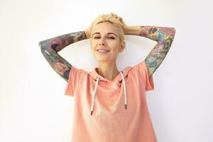 Cheerful young pretty tattooed woman holding her blonde hair with raised hands and looking positively at camera with charming smile, isolated over white background photo