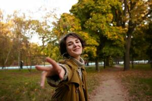 Positive young attractive brunette lady with casual hairstyle smiling pleasantly at camera and reaching out raised hand, walking over yellowed trees in city park photo