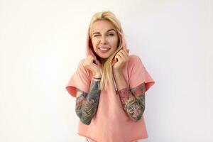 Indoor shot of young green-eyed tattooed blonde female keeping one eye closed while looking positively at camera and raising hands to her face, isolated over white background photo