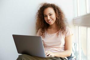 Portrait of positive young curly african american girl sitting by the window, dressed in blue pajamas, smiling and enjoying his favorite video at freelance work. photo