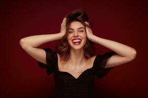 Indoor shot of young charming brunette lady in elegant black top with red dots clutching happily her head and laughing cheerfully while posing over claret background photo