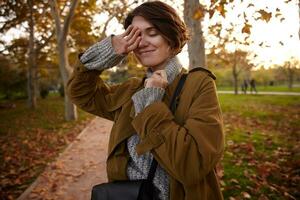 Good looking young positive short haired brunette woman raising hand to her face and smiling pleasantly with closed eyes while standing over park background photo