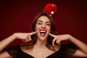 Christmasnew year themed portrait of young cheerful female with brown wavy hair holding forefingers on cheeks and smiling cheerfully at camera, celebrating holiday with friends over claret background photo