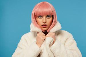 Attractive young pink haired female with bob haircut wearing colored makeup while posing over blue background, holding hand on collar of her white coat and looking at camera photo