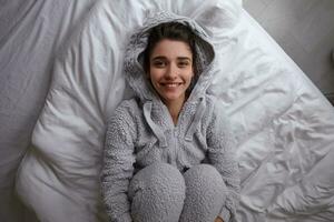 Indoor shot of happy attractive young brunette female dressed in cosy clothes lying on white bed linens and looking positively at camera with wide smile, holding legs with her hands photo