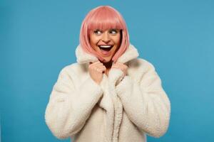 Overjoyed attractive young female with short pink bob haircut looking exitedly aside with wide eyes and mouth opened, holding colar of her white coat while posing over blue background photo