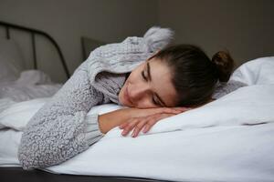 Close-up of sleepy pretty young brunette woman with bun hairstyle keeping head on folded hands and closing her eyes, posing over bedroom interior in home cosy clothes photo