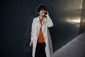 Good looking smiling young curly brunette lady having pleasant conversation on phone while posing over black urban wall, wearing stylish clothes and wide grey hat photo