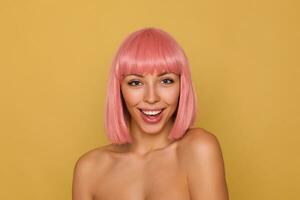 Indoor photo of beautiful young blue-eyed pink haired woman with bob haircut looking joyfully at camera and smiling widely while standing over mustard background