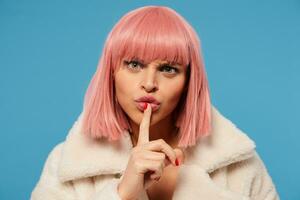 Close-up of young green-eyed pretty pink haired female holding forefinger on her lips and frowning eyebrows while looking seriously at camera, isolated over blue background photo